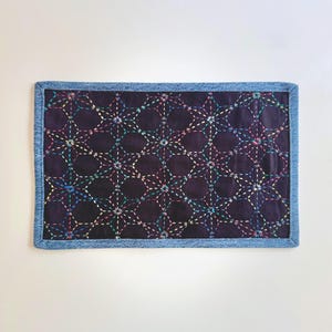 May include: A rectangular fabric mat featuring a dark purple center panel with a repeating geometric star pattern stitched in multicolored thread. The mat is finished with a light blue denim fabric border around all four edges.