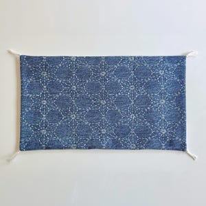 May include: A rectangular denim fabric piece featuring a white stitched geometric star pattern across the surface. The blue textile has frayed white thread tassels at each of the four corners, creating a textured and detailed appearance.