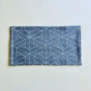 May include: A rectangular, slate blue fabric item with a geometric pattern of white stitching. The stitching forms a repeating pattern of interconnected shapes, creating a textured surface. The item is likely a decorative piece or a small textile.