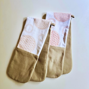 May include: Two beige oven mitts with white tops featuring embroidered orange circles and floral patterns. The mitts have a loop for hanging and are made of a soft, durable fabric. These kitchen accessories are designed for heat protection while handling hot cookware.