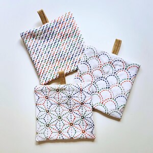 May include: Three white fabric pot holders with decorative stitching and brown leather hanging loops. One has a zig-zag pattern in red, green, and blue. Another features a scale-like design, and the third has a starburst pattern.