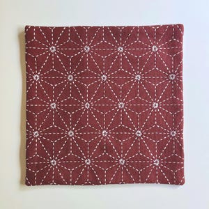 May include: A square fabric cushion cover in a deep maroon color featuring a white stitched geometric pattern. The design consists of repeating star-like shapes formed by intersecting lines, creating a consistent grid across the entire surface.