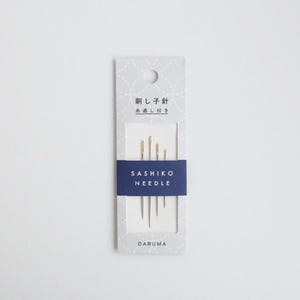 May include: A package of Sashiko needles, featuring a blue band with white text. The package has Japanese text at the top and the brand name "DARUMA" at the bottom. The needles are gold and silver in color.