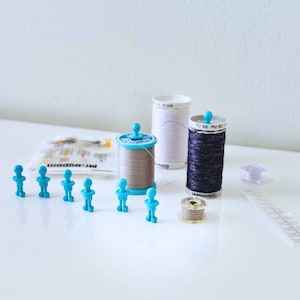 May include: A collection of sewing supplies, including spools of thread in various colors, a bobbin, and small blue thread holders shaped like people. The spools are in shades of gray, white, and navy blue. A package of pins is also visible.