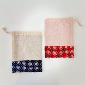 May include: Two drawstring bags. One is light pink with a red patterned bottom, the other is beige with a navy blue patterned bottom. Both bags have a natural-colored drawstring closure at the top. The bags are made of a natural fabric.