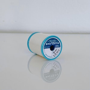 May include: A white spool of thread with blue lettering that reads "クラフト".  The spool is labeled "シャッペスパン" and "200m 100% Polyester".  The spool also has a label that reads "TOHOBU 生糸 403".