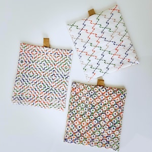 May include: Three square fabric items with colorful embroidered patterns. Each has a different design, including geometric and floral motifs, in shades of orange, green, blue, and white. They have a small, tan leather loop at the top.