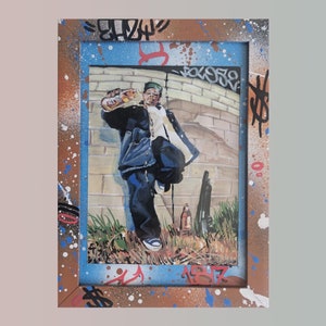May include: A framed painting of a person wearing a blue jacket and jeans, holding a bottle in their hand. The painting is set against a backdrop of a brick wall with graffiti.