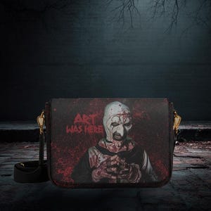 May include: Black crossbody bag with a horror-themed graphic. The image on the bag shows a character with a white mask and blood-like splatters, with the text "ART WAS HERE". The bag has a black strap and gold-coloured hardware.