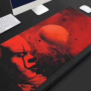May include: A rectangular desk mat featuring a high-contrast red and black graphic illustration. The design depicts the profile of a clown with a prominent forehead and dark eye makeup, positioned next to a single floating balloon in the center.