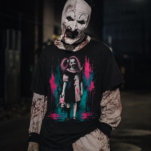 May include: A black t-shirt featuring a graphic print of a dark-haired girl in a clown costume holding a doll, surrounded by pink and teal paint splatters. The shirt is worn over a long-sleeved garment with blood-like stains and a clown mask.
