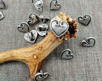 May include: A collection of silver-colored heart-shaped buttons with decorative swirls. The buttons are scattered on a textured gray surface, with some resting on a piece of antler. The buttons have a vintage or antique appearance.