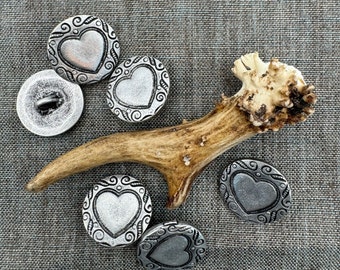 May include: Five silver-toned metal buttons with a heart design and ornate detailing. The buttons are arranged on a gray fabric background with a piece of antler bone.