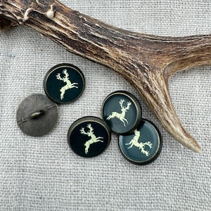 May include: Five round, dark green buttons with a gold deer silhouette design. The buttons are arranged in a semi-circle on a light brown, textured surface.