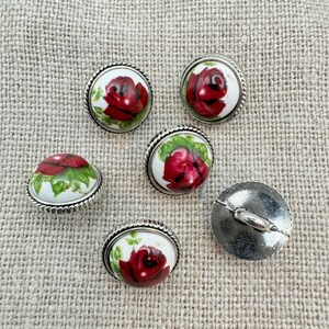 May include: Six round, silver-rimmed buttons with a white background and a red rose design. One button is shown from the back, revealing a silver loop.