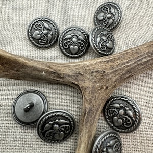 May include: A set of eight silver-toned metal buttons with a floral design featuring a heart and birds. The buttons are arranged on a piece of antler.