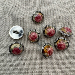 May include: Assortment of eight round, silver-rimmed buttons with a floral design. The buttons feature a cream-colored background with pink, yellow, and blue flowers.