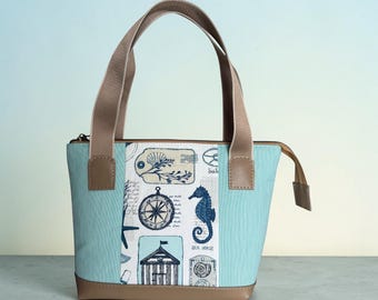 May include: A tote bag featuring a central white panel with navy blue nautical illustrations including a compass, a seahorse, a beach hut, and script text. The sides are light blue, and the base and handles are a solid tan color with a zipper.