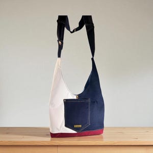 May include: A denim shoulder bag with a color block design. The bag features a white panel, a navy blue panel, and a navy blue pocket. The bottom of the bag is trimmed with a maroon band. The bag has a long, adjustable strap.