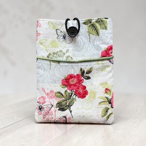 May include: A rectangular fabric pouch featuring a floral and butterfly print in shades of red, green, and grey on a white background. The pouch includes a front pocket, a green piped edge, and a black elastic cord closure with a single black button.