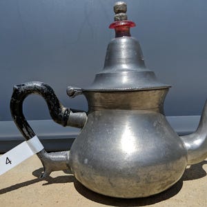 Könnte beinhalten: Eine silberfarbene Vintage-Teekanne mit einem gebogenen schwarzen Griff und einem roten und klaren Glasverschluss. Die Teekanne hat einen abgerundeten Körper, einen Ausguss und einen Deckel. Ein weißes Etikett mit der Nummer "4" ist am Griff befestigt.