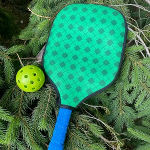 Pu&ograve; includere: Una racchetta da pickleball verde con un'impugnatura blu e una palla da pickleball verde su un letto di aghi di pino verdi.