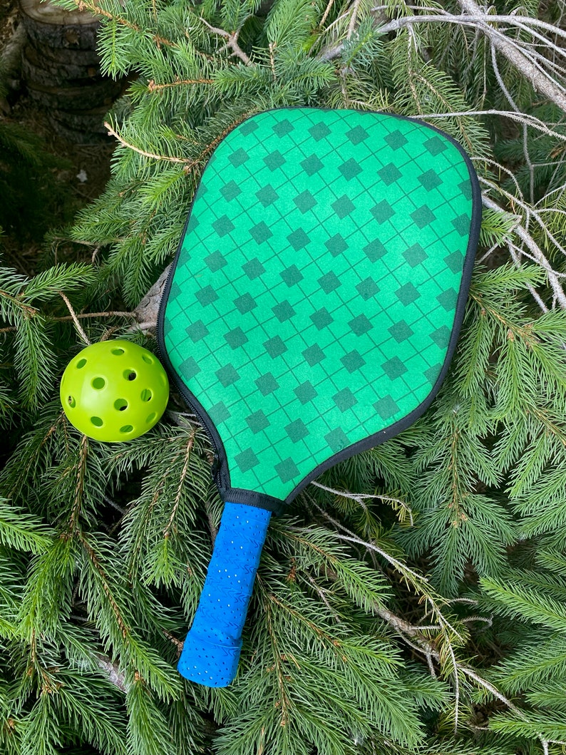 Pu&ograve; includere: Una racchetta da pickleball verde con un'impugnatura blu e una palla da pickleball verde su un letto di aghi di pino verdi.