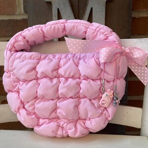 May include: A pink quilted handbag with a rounded shape and a padded handle. The bag features a pink polka dot ribbon tied in a bow. A small charm with a cartoon character is attached to the side of the bag.