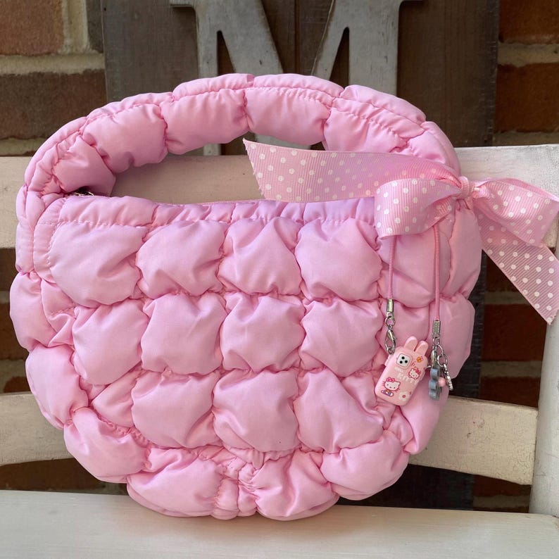 May include: A pink quilted handbag with a rounded shape and a padded handle. The bag features a pink polka dot ribbon tied in a bow. A small charm with a cartoon character is attached to the side of the bag.