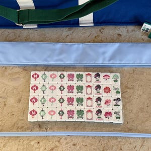 May include: A set of Mahjong tiles with various designs, including lanterns, flowers, and a woman's face, are displayed inside a clear, light blue zippered case. A blue tote bag with a green strap is visible in the background, along with a few green and white tiles.