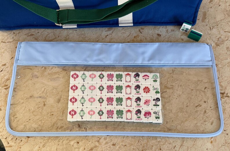 May include: A set of Mahjong tiles with various designs, including lanterns, flowers, and a woman's face, are displayed inside a clear, light blue zippered case. A blue tote bag with a green strap is visible in the background, along with a few green and white tiles.