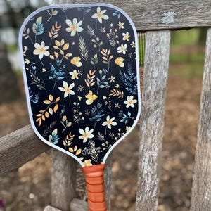 May include: A black and white paddle with a floral pattern featuring yellow, white, and blue flowers and leaves. The paddle has an orange handle and the text "CHELSEA MEL" is printed on the paddle.