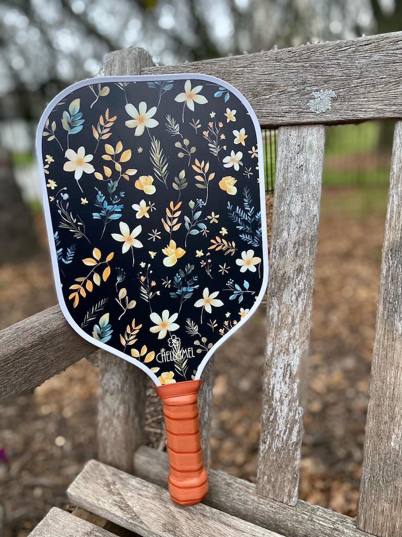 K&ouml;nnte beinhalten: Ein schwarz-wei&szlig;es Paddel mit einem Blumenmuster mit gelben, wei&szlig;en und blauen Blumen und Bl&auml;ttern. Das Paddel hat einen orangefarbenen Griff und der Text "CHELSEA MEL" ist auf dem Paddel aufgedruckt.