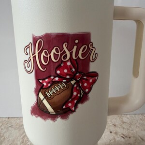 May include: Cream-colored insulated tumbler with a handle and a copper-colored rim. The tumbler features a graphic of a football, a red polka-dot bow, and the word "Hoosier" in a script font, set against a maroon background.