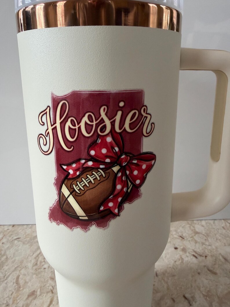 May include: Cream-colored insulated tumbler with a handle and a copper-colored rim. The tumbler features a graphic of a football, a red polka-dot bow, and the word "Hoosier" in a script font, set against a maroon background.