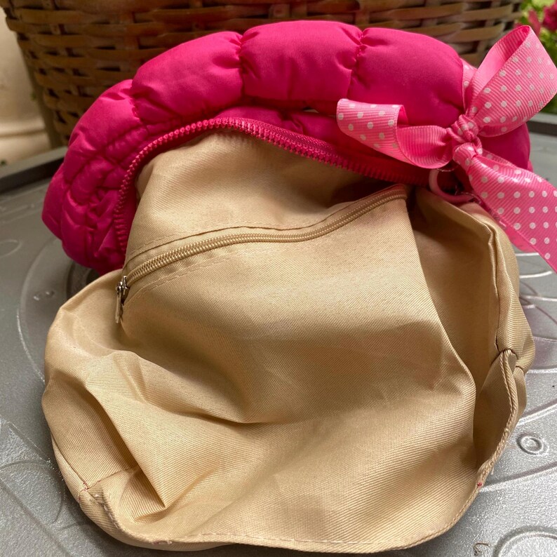 May include: A quilted, hot pink bag with a beige interior. The bag has a zipper closure and a pink bow with white polka dots. The bag is open, revealing the interior lining.