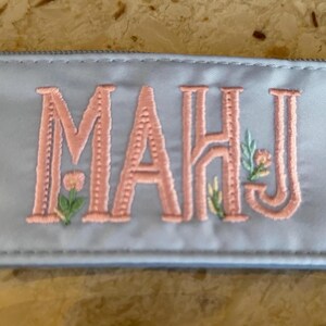 May include: A light blue pencil case with the embroidered word "MAHJ" in a coral color. The letters are embellished with floral designs in green and yellow. The case is made of a smooth, satin-like material.