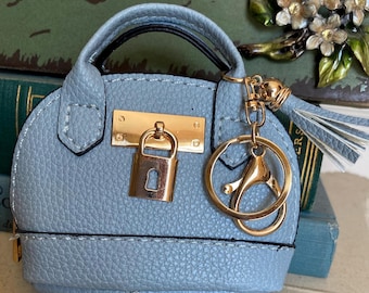 May include: A light blue miniature handbag keychain with a gold-colored lock and hardware. The small purse has a textured surface, a black handle, and a matching tassel. The keychain is designed to resemble a classic handbag.