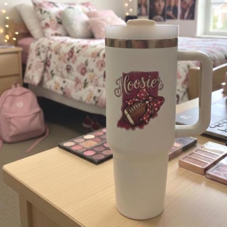 May include: A white insulated tumbler with a handle and a gold band near the top. The tumbler features a graphic of a football, a bow, and the word "Hoosier" in a script font. The tumbler is on a wooden table.