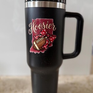 May include: Black travel mug with a handle and a black lid. The mug features a graphic of the state of Indiana with the word "Hoosier" in cursive, a football, and a red polka dot bow.