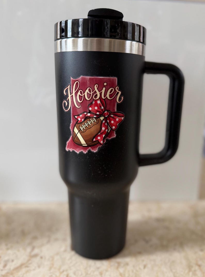 May include: Black travel mug with a handle and a black lid. The mug features a graphic of the state of Indiana with the word "Hoosier" in cursive, a football, and a red polka dot bow.