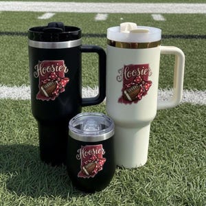 May include: Three insulated tumblers in black and cream colors, and a black wine tumbler. Each tumbler features the word "Hoosier" with a football and bow graphic. The tumblers have handles and lids, perfect for hot or cold beverages.