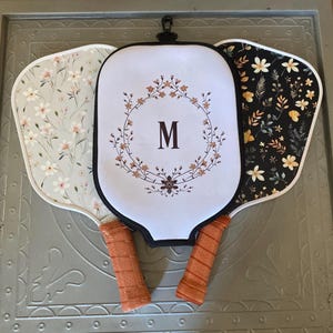 May include: Three white pickleball paddles with black trim. The paddles have different designs. One has a floral pattern, one has a floral wreath with the letter M in the center, and one has a black and white floral pattern.