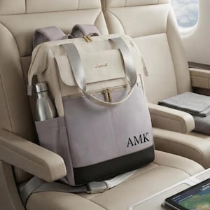 May include: A multi-toned backpack featuring a cream-colored top flap with gold-tone hardware, a light gray main body with the black embroidered letters AMK, and a black base. A silver metal water bottle is tucked into the side pocket.