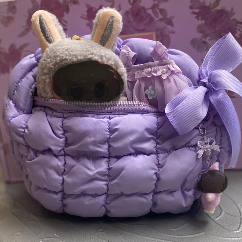 May include: A quilted lavender bag with a matching bow and charm. Inside, a plush bunny hat and a small lavender dress are visible. The bag is designed with a zipper closure and a decorative bow, suggesting a gift or storage item.