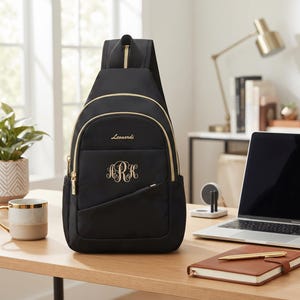May include: A black sling backpack featuring gold-tone zippers and hardware. The front displays the name Leonardi in gold script above a decorative three-letter monogram in gold embroidery. The bag has a single shoulder strap and a front pocket.