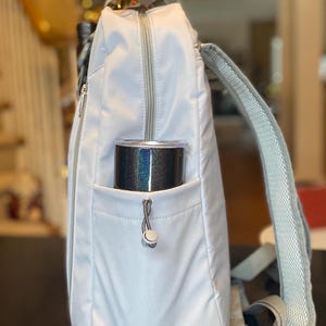 Op de afbeelding: Een lichtgrijze rugzak met een zilveren rits en bandjes. Een zwarte en iriserende beker zit in de zijzak. De rugzak heeft een handvat aan de bovenkant en verstelbare bandjes.