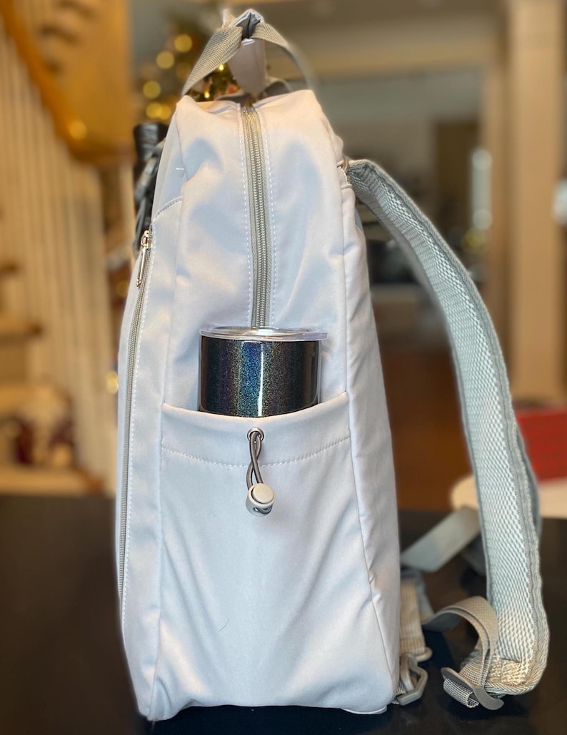 Op de afbeelding: Een lichtgrijze rugzak met een zilveren rits en bandjes. Een zwarte en iriserende beker zit in de zijzak. De rugzak heeft een handvat aan de bovenkant en verstelbare bandjes.