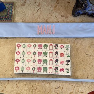 May include: A light blue zippered pouch with the embroidered letters "MAHJ" in pink. Inside the clear pouch are Mahjong tiles with various symbols and characters. A small blue bag with gold trim and the word "Mahjong" is in the background.