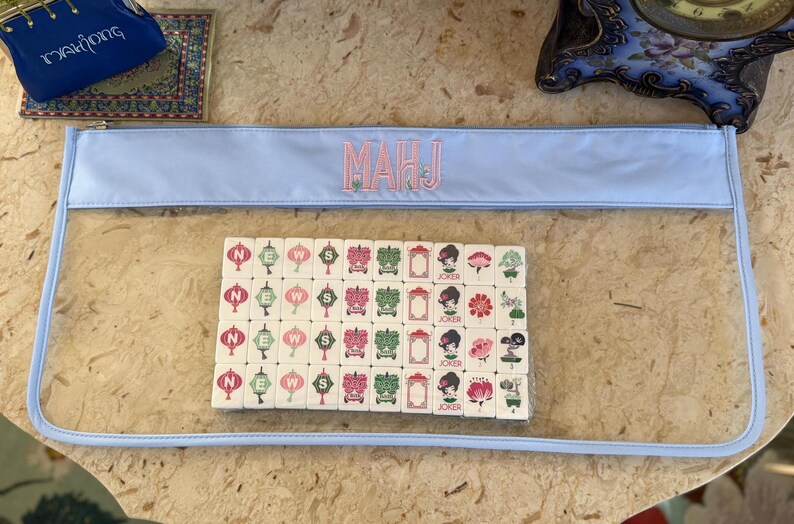 May include: A light blue zippered pouch with the embroidered letters "MAHJ" in pink. Inside the clear pouch are Mahjong tiles with various symbols and characters. A small blue bag with gold trim and the word "Mahjong" is in the background.